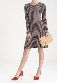 Femme portant une robe à manches longues rayée, tenant une pochette beige et chaussant des escarpins rouges, se tenant devant un fond uni.