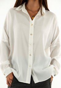 Camisa blanca de mangas largas en tejido ligero, con cierre de botones en el frente con botones transparentes y crema, y un ajuste relajado con puños enrollados.