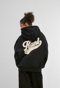 Zwarte hoodie met een groot crèmekleurig logo "Kani" op de achterkant. Beschikt over een capuchon met trekkoord en een ontspannen pasvorm. Gemaakt van zachte stof.