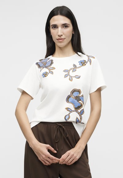 Γυναίκα που φοράει λευκό κοντομάνικο μπλουζάκι με μπλε και καφέ floral σχέδια, συνδυασμένο με καφέ παντελόνι με κορδόνι, στέκεται μπροστά σε απλό φόντο.