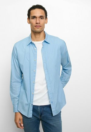 Hellblaues Hemd mit Knöpfen und Kragen, aus einem strukturierten Stoff, über einem weißen T-Shirt getragen. Lange Ärmel und lässiger Stil.