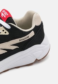 Black and beige sneaker featuring mesh and suede materials, white midsole, brown rubber outsole, and red branding on the tongue.