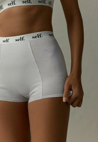 Valkoiset puuvillaiset hipster-shortsit mustalla brändäväellä. Kangas on sileä, joustava ja istuva, korostaen jalan muotoa.