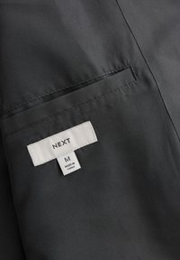 Giacca nera in tessuto con una tasca e un’etichetta con la scritta "NEXT", taglia "M" e "FABBRICATO IN CINA". Texture liscia, design minimalista.