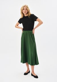 Czarna koszulka z krótkim rękawem zestawiona z plisowaną zieloną spódnicą do kolan. Spódnica ma zgromadzony pas i gładką teksturę. Czarne baleriny dopełniają stylizację.