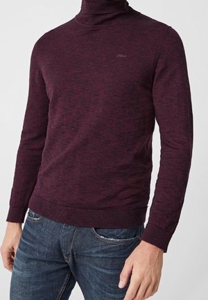 Homme portant un pull col roulé ajusté bordeaux associé à un jean en denim bleu sur un fond uni.