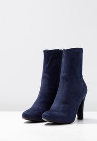 Par de botas de ante azul marino hasta el tobillo con cremallera lateral y tacones altos en una superficie blanca con un fondo gris claro.