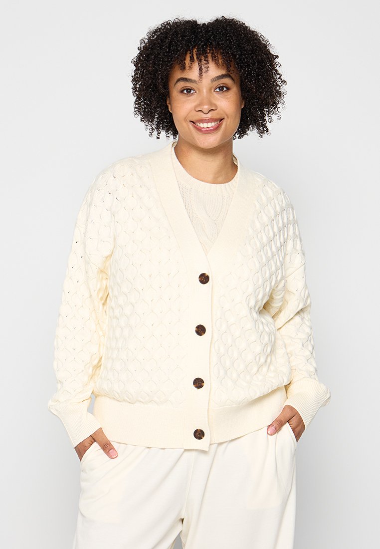 Crèmekleurige gebreide cardigan met ruitpatronen, houten knopen, geribde manchetten en zoom, gedragen over een lichte trui.