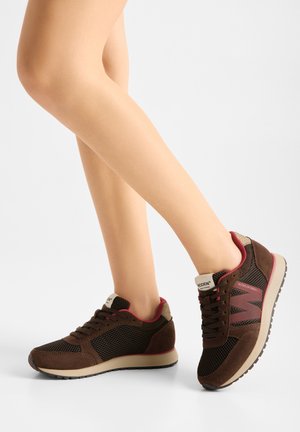 Beine, die braune und schwarze Mesh-Sneakers mit roten Akzenten und beigen Sohlen tragen, stehen mit einem leicht angehobenen Fuß auf weißem Hintergrund.
