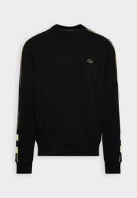 Svart sweatshirt med långa ärmar, med gula ränder längs sidorna och en liten grön logotyp på bröstet. Mjuk textur.