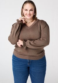 Bruine, glinsterende trui met lange mouwen, V-hals en opgeblazen mouwen; gecombineerd met blauwe spijkerbroek. Met gerimpelde manchetten.