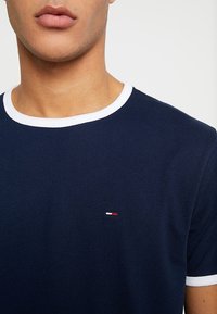 Navy blue cotton t-shirt with white ribbed collar and sleeve hems. Features a small, embroidered logo in red, white, and blue on the chest.