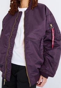Lila nylon bomberjacka med en gulddragkedja, ribbade ärmslut och fickdetaljer. Har en röd etikett med texten "REMOVE BEFORE FLIGHT."