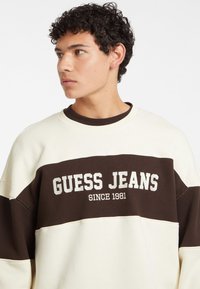 Sudadera en crema claro y marrón con el texto "GUESS JEANS", que presenta un corte relajado, cuello redondo y bloques de color contrastantes.