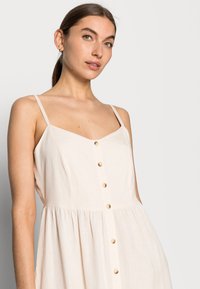 Hellbeige ärmelloses Kleid mit V-Ausschnitt, Frontknöpfen und gerafftem Bund. Hergestellt aus einem strukturierten Stoff mit lockerer Passform.