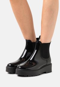 Bottines Chelsea en vernis noir avec panneaux élastiques sur les côtés, semelle basse à plateforme et semelle extérieure en caoutchouc texturé. Fini lisse avec un design à bout arrondi.
