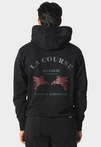 Persona con sudadera negra con capucha levantada, que presenta diseños de caballos alados rojos y texto en francés en la parte posterior.