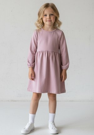 Młoda dziewczyna o blond włosach, ubrana w różową sukienkę z długim rękawem, białe skarpetki i białe trampki, stoi na tle jednolitego jasnego tła.