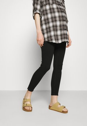 Person iført sorte leggings, en brun ternet skjorte og brune sandaler med dobbelt rem, stående mod en ensfarvet hvid baggrund.