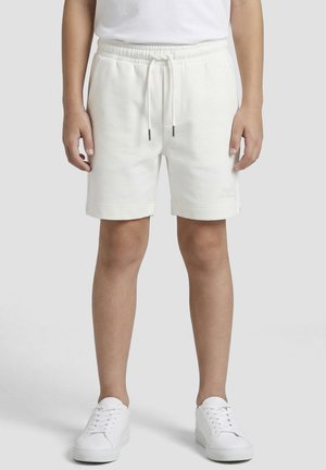 Enfant portant un short blanc uni avec cordon de serrage et des baskets blanches, debout devant un fond neutre.