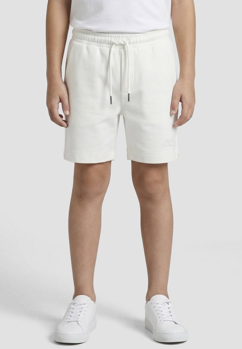 Enfant portant un short blanc uni avec cordon de serrage et des baskets blanches, debout devant un fond neutre.