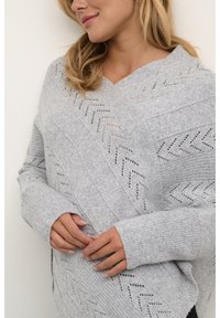 Pelēks adīts džemperis ar V-veida kaklu, ar zigzaga rakstu un ribotām piedurknēm. Mīksta tekstūra un brīva piegriezuma, kas nodrošina ērtu valkāšanu.