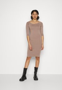 Vestido justo de manga longa em tecido castanho claro, até aos joelhos, com decote redondo. Acompanhado de botas de tornozelo pretas. Design simples.