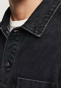 Camisa de mezclilla negra con cuello puntiagudo, bolsillo en el pecho y cierre de botones. Presenta una tela texturizada y detalles de costuras visibles.