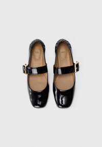 Par de zapatos Mary Jane de charol negro con punteras cuadradas y correas de hebilla dorada sobre un fondo claro.