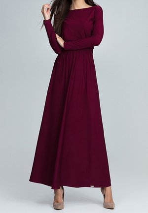 Femme portant une robe longue rouge foncé à manches longues, taille cintrée et talons hauts beiges sur un fond uni.