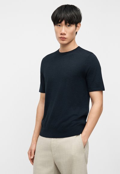 Νεαρός άνδρας με κοντά μαύρα μαλλιά, ντυμένος με εφαρμοστό μαύρο πουλόβερ κοντών μανικιών και μπεζ παντελόνια, στέκεται με το ένα χέρι στην τσέπη.