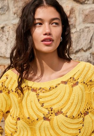 Hullámos sötét hajú nő karika fülbevalóval és sárga, banánmintás kötött felsőben, egy kőfal előtt állva.