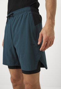 Pantalones cortos deportivos para hombres en color teal con una banda negra en la cintura, que cuentan con un cordón ajustable, bolsillos laterales y un forro interior corto para mayor comodidad.