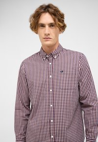 Młody mężczyzna z kręconymi włosami, ubrany w koszulę w kratę w czerwono-białe-niebieskie kolory, na tle jasnego, jednolitego tła.