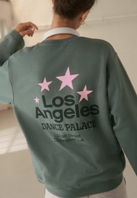Sudadera verde con "Los Ángeles" en texto negro y "DANCE PALACE" debajo, acentuado con tres estrellas rosas. Material suave, corte relajado.