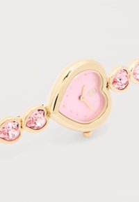 Χρυσό ρολόι βραχιόλι με ροζ καντράν σε σχήμα καρδιάς και αντίστοιχους ροζ καρδιές από πολύτιμους λίθους στα συνδετικά του αλυσίδας, δείχνοντας ώρα 4:40.