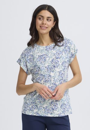 Kobieta ubrana w biały krótki rękaw z małym niebiesko-zielonym kwiatowym nadrukiem, w połączeniu z ciemnoniebieskimi spodniami, spoglądająca na bok.