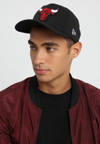 Gorra de béisbol negra con un logotipo de toro bordado en rojo y blanco en la parte frontal, llevada por una persona con una chaqueta bomber burdeos sobre una camisa negra.
