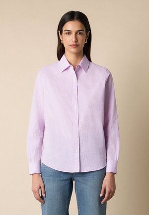 Młoda kobieta o prostych ciemnych włosach nosi jasnofioletową kraciastą koszulę na guziki i niebieskie dżinsy, stoi na beżowym tle.