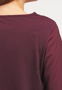 Burgunderfarbenes Langarmoberteil aus einem weichen, leichten Stoff. Besitzt einen einfachen Rundhalsausschnitt und eine glatte Textur, von hinten gesehen.