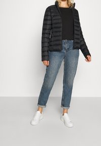 Svart puffjacka med horisontellt quiltat mönster, hög krage, bärs över en svart topp. Kombinerad med ljusblå jeans med uppvikta fållar och vita sneakers.