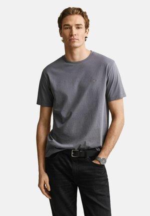 Junger Mann trägt ein schlichtes graues T-Shirt mit kurzen Ärmeln, schwarze Hose, schwarzer Gürtel und silberne Armbanduhr, steht mit einer Hand in der Tasche.