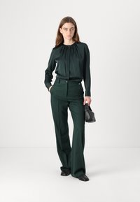 Kvinna i mörkgrön blus och byxor som håller en svart handväska, står mot en enkel vit bakgrund.
