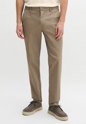 Mann trägt beige gerade geschnittene Hosen und graue Wildleder-Schnürschuhe und steht vor weißem Hintergrund.