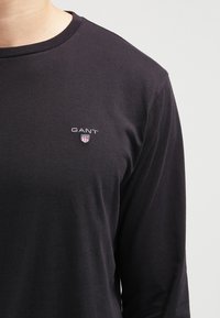 Musta pitkähihainen T-paita, joka on valmistettu puuvillasta. Paidassa on pyöreä kaula-aukko ja pieni brodattu logo rinnassa valkoisilla ja punaisilla yksityiskohdilla.