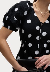 Schwarze Bluse mit Polka-Dots, Puffärmeln, strukturiertem Stoff, V-Ausschnitt und Zugbanddetail. Das Modell hält eine schwarze Handtasche.