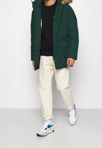 Giacca parka verde con cappuccio foderato in pelliccia, dotata di tasche frontali, maglione nero sotto, pantaloni crema e sneakers bianche con accenti blu.