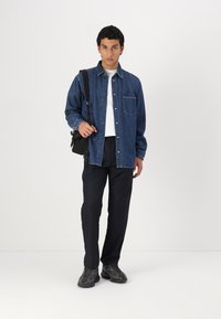 Camisa de mezclilla de botones en azul oscuro, de manga larga, con un bolsillo frontal. Lleva puesta sobre una camiseta blanca, combinada con pantalones navy y zapatillas negras.