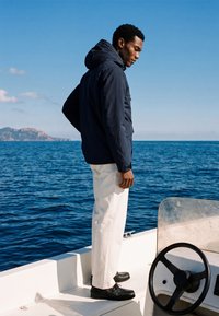 Homme en veste sombre, pantalon blanc et chaussures noires, debout sur le pont d'un bateau, regardant l'eau avec des montagnes en arrière-plan.