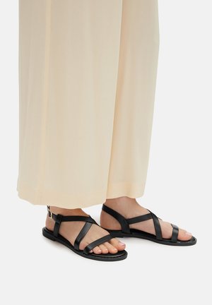 Czarne skórzane sandały z krzyżującymi się paskami i płaską podeszwą, noszone z jasnymi, szerokimi spodniami, podkreślające minimalistyczny design.